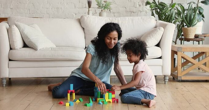 Young Afro American mother play constructor on warm heated floor with preschool age daughter child enjoy building castle from wooden details. Bonding mom kid relax create toy constructions from cubes