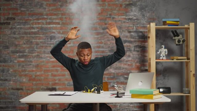 Funny African-American guy technician with smoke rising above head scared of short circuit shakes hands at workplace in scientific laboratory