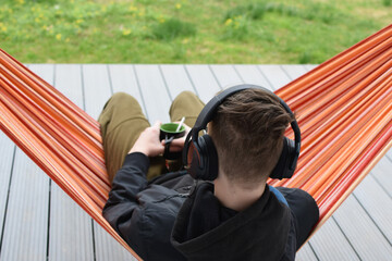Young male person enjoys listening music in headset from cell phone and resting in hammock outdoors. Back view.
