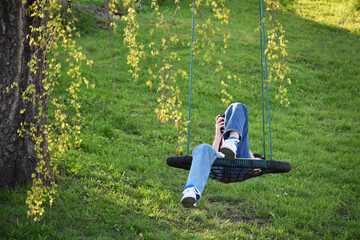 Young teen girl texting phone and resting in nest swing outdoors in summer day. Green grass background.
