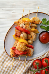 Plate of grilled chicken skewers with vegetables and sauce on white wooden background