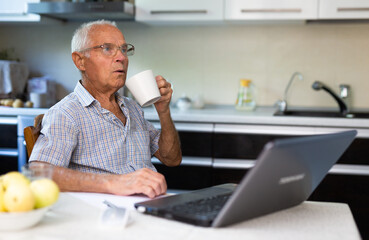 Old man sitting in kitchen at table with laptop