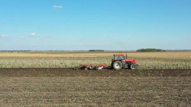 Low place of view with dolly move shot of tractor as pulling machine, harrowing arable field, preparing soil for planting new crop next season.