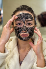 the girl puts a black mask on her face made of black clay and corrects it to the mirror.