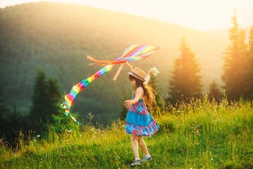 Little girl playing with kite. The child is dressed in a summer dress and hat. Mountain landscape on background. Sunny summer day.
