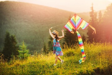 Little girl playing with kite. The child is dressed in a summer dress and hat. Mountain landscape on background. Sunny summer day.