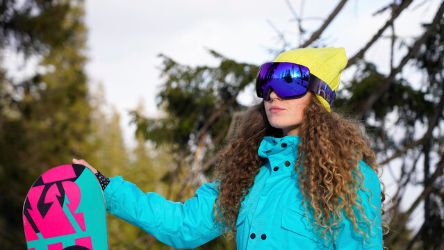 Bright curly girl snowboarder in a yellow-blue suit against the backdrop of a mountain forest.