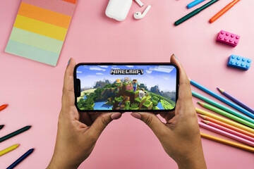 Naklejka premium Girl holding a smartphone with Minecraft mobile game app on the screen. Pink background with school supplies, accessories, video game controller. Rio de Janeiro, RJ, Brazil. March 2022