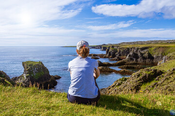 Woman sitting on the edge of a cliff in Arnarstapi, relaxing and thinking while admiring the landscape. Snaefellsnes peninsula, Iceland
