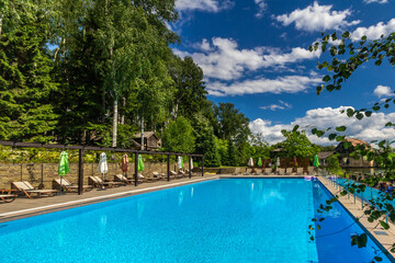 Large outdoor swimming pool with sun beds and umbrellas on a background of trees, on a clear day.