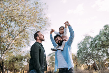 Gay couple with their young daughter enjoying a day in a park. The father raises the arms of the...