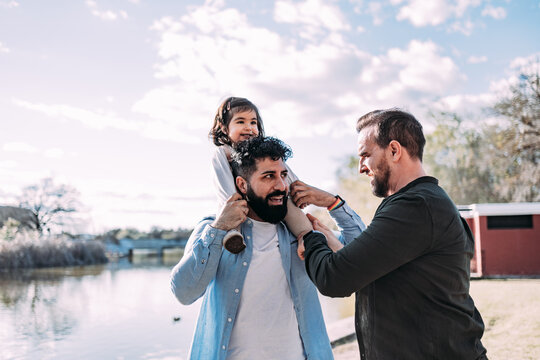 Gay couple with their little daughter on the shore of a lake in the park. The father holds the girl on his shoulders piggyback.