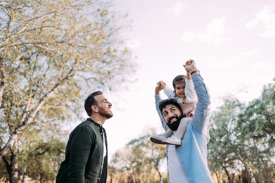 Gay couple with their little daughter in the park. The father holds the girl on his shoulders in a piggyback ride and they raise their arms happily.
