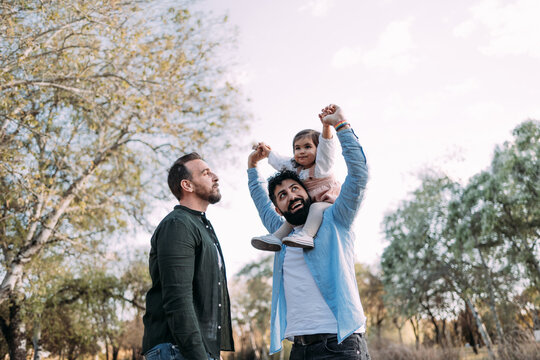 Gay couple with their young daughter enjoying a day in a park. The father raises the arms of the daughter smiling at her.