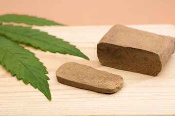 hashish tablet, hashish portion 100 grams, with large marijuana leaf, on wood, brown background - obrazy, fototapety, plakaty