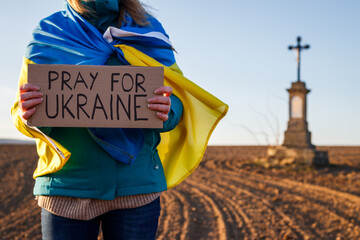 Pray for Ukraine. Woman wrapped in ukrainian flag holding anti war sign cardboard outdoors....