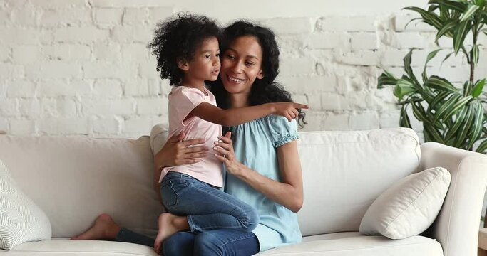 Loving Black single mum sit on comfy couch with adorable preschool age daughter rest talk cuddle give affectionate kisses one another. Kind foster mom hug beloved little girl chat have fun with child