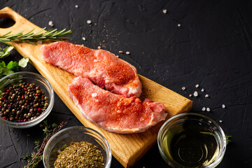 Raw meat on a cutting board. Dark background. Preparation for cooking pork meat. Various spices, seasonings lie nearby.