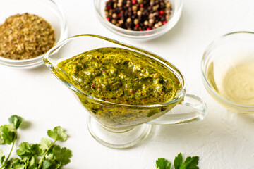 Chimichurri sauce in a gravy bowl on a white background. Various spices lie nearby. Argentinean vegetarian sauce made from olive oil, oregano, parsley.