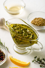 Chimichurri sauce in a gravy bowl on a white background. Various spices lie nearby. Argentinean vegetarian sauce made from olive oil, oregano, parsley.