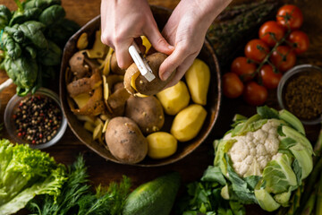 A woman is peeling potatoes. Various fresh vegetables lie on the table. Cooking vegetarian food.