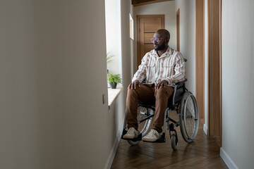 Young African American man with disability looking through window while sitting on wheelchair in...