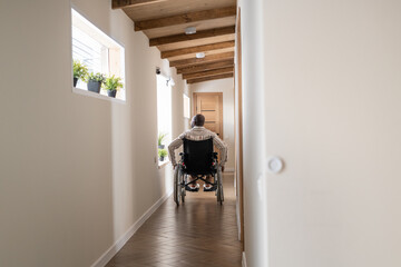 Back view of man with paralysis moving on wheelchair along corridor of large cotemporary apartment...