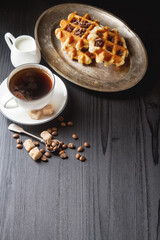 Homemade belgian waffles, white ceramic cup of coffee, milk, teaspoon and coffee beans. Dark rustic...