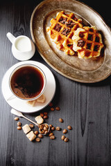 Homemade belgian waffles, white ceramic cup of coffee, milk, teaspoon and coffee beans. Dark rustic...