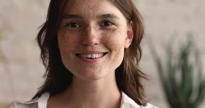 Close up portrait beautiful cheerful female teenager looking at camera then aside with white sincere smile on pretty face full of freckles. Happy joyful young attractive red haired lady posing indoors