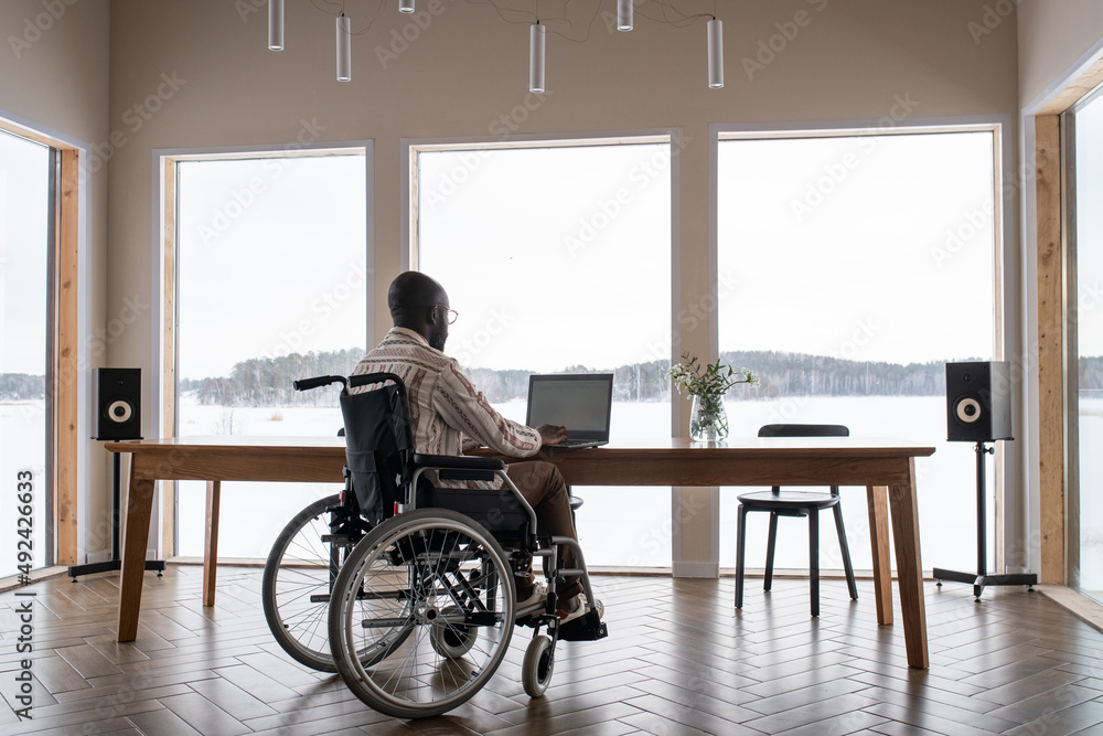 Poster African American man with disability working in front of laptop while sitting by table in front of lapge window in living room