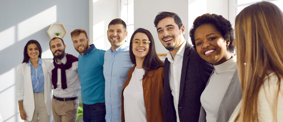 Team of happy united young business people hugging each other. Diverse mixed race group of men and women standing in row in their office, huddling and smiling all together. Unity and teamwork concept