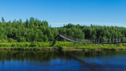 Wooden pedestrian suspension old bridge over a river, over forest and pond. North Siberia.Russia