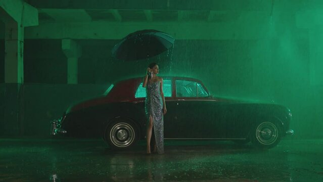 Beautiful Elegant Young Woman Standing and holding umbrella near old retro classic car. Girl with umbrella stands in street under the rain . Old fashioned luxury style concept . Autumn rainy day
