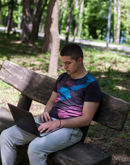 A young handsome man is sitting on a wooden bench and working on his laptop in nature.