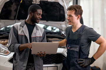 Computer Car Tuning. two Smiling auto mechanic testing a car engine connected to laptop. in auto repair shop. diverse men in uniform having conversation, discussion. cooperation, team work