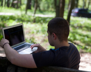 A young handsome man is sitting on a wooden bench and working on his laptop in nature.
