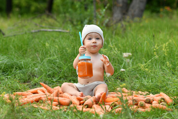A little boy, one 1 year old, holding a jar of carrot juice on a pile of carrots in nature near a forest in a clearing and holds carrots in his hands.