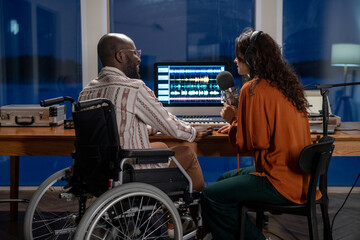 Young African American man in wheelchair looking at female in headphones singing in microphone...