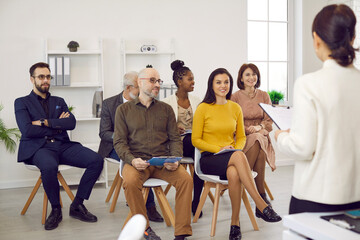 Female business coach in meeting in office speaks in front of small group of business people. Group of multiracial diverse contented people sitting on chairs attentively listening to their leader. - obrazy, fototapety, plakaty