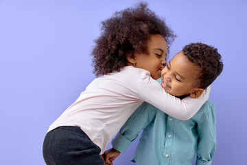 Two cheerful black american siblings, sister kissing her brother, isolated on purple background....