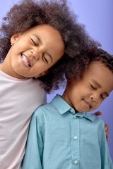 Obraz premium Two happy black children posing at camera and cheerfully smiling over purple studio background, cute american brother and sister having fun together, wearing casual shirts. childhood concept