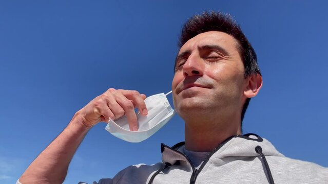 Young man removes ffp2 protection mask and breathes with smile under blue sky. Male person soothed by end of pandemic concept