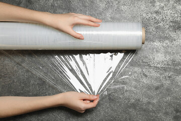 Woman unrolling transparent plastic stretch wrap on grey stone background, top view