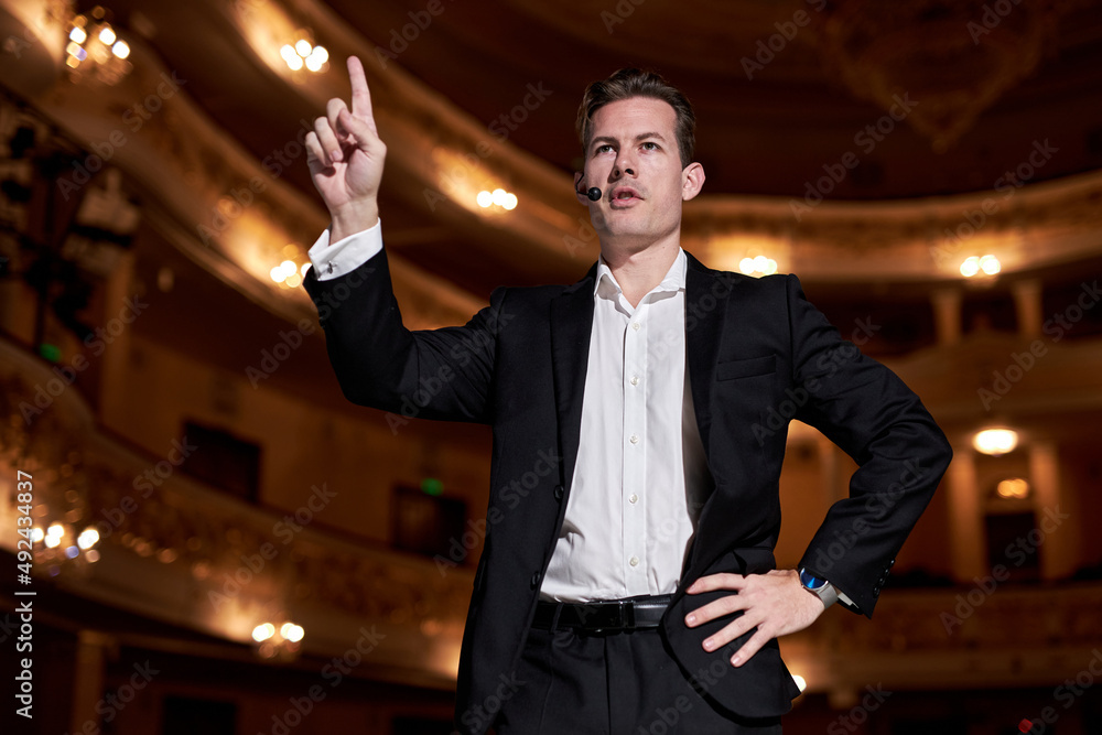 Wall mural male in suit gesticulating and speaking to audience while standing on stage in theater. male speaker is doing the public speaking, motivational speaking, inspiration speaker concept