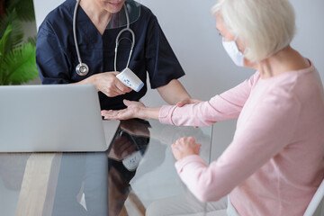Female doctor in transparent visor measuring temperature of elderly woman in mask