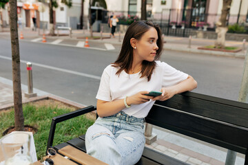 Fototapeta na wymiar Stylish attractive woman in white t-shirt and jeans is sitting on the bench with smartphone and looking aside in the city in warm spring day