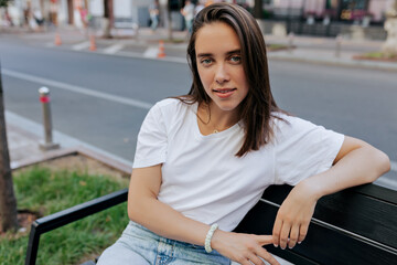 Young cute stylish girl is resting on the bench in warm spring day during lunch. Pretty student is sitting in a open-air cafe near the brick building