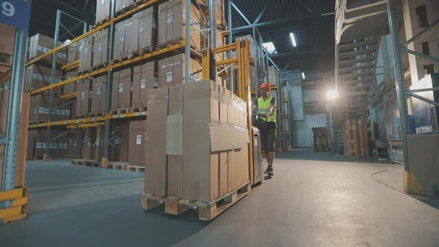A worker in a warehouse carries a Pallet Stacker Truck with goods. Worker with rocklay in the warehouse. The worker walks through the warehouse