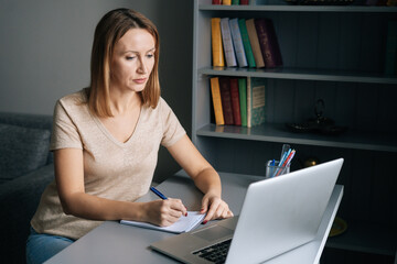 High-angle view of serious woman write making notes, studying online on laptop at home looking to screen. Attractive female take remote web training or course on laptop, talking on video call.